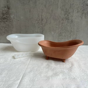Op de afbeelding: Twee witte keramische badkuipen, een groter dan de andere, met een kleine witte plastic mal met zes gaten. De kleinere badkuip is terracotta gekleurd en heeft vier kleine pootjes.