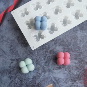 Puede incluir: Un molde de silicona blanco con 18 cavidades en forma de flor. Se muestran tres golosinas terminadas, de cuatro lóbulos, con forma de caramelo en azul, verde y rosa.