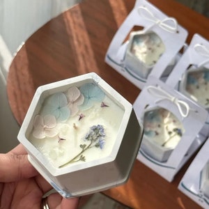 Puede incluir: Una vela hexagonal blanca con flores secas incrustadas en la cera. La vela está en un recipiente blanco con un diseño de mármol.