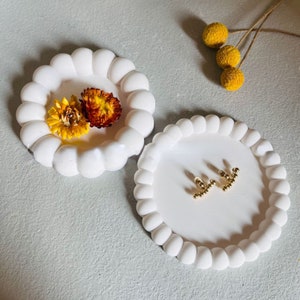 Può includere: Due piatti in ceramica bianca con bordi a festoni. I piatti sono perfetti per contenere piccoli oggetti come gioielli o cianfrusaglie. Un piatto contiene fiori secchi, mentre l'altro contiene un paio di orecchini d'oro.