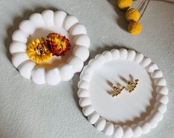 May include: Two white ceramic dishes with scalloped edges. The dishes are perfect for holding small items like jewelry or trinkets. One dish has dried flowers in it, while the other has a pair of gold earrings.