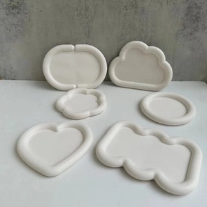 May include: Six white ceramic trays in different shapes, including a heart, a cloud, a circle, and an oval. The trays are all smooth and have a slightly raised edge.