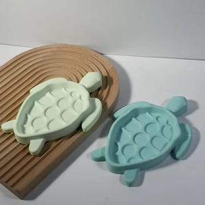 May include: Two turtle-shaped soap dishes, one light green and one teal, made of silicone. The dishes are on a wooden surface.