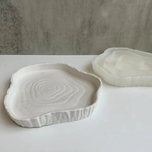 May include: Two white ceramic trays with a tree ring design. The tray on the left has a smooth surface and the tray on the right has a raised edge.