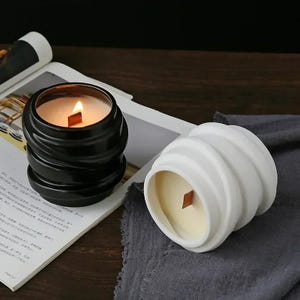 Puede incluir: Dos velas decorativas en recipientes de cerámica negra y blanca con una mecha de madera. Las velas tienen un diseño ondulado y texturizado.