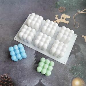 Puede incluir: Un molde de silicona blanco con seis cavidades cuadradas, cada una con un diseño de burbujas en relieve. Se muestran tres barras de jabón terminadas: una azul, una verde y una blanca.