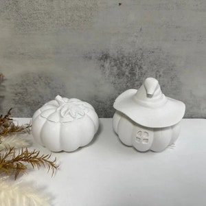 May include: Two white ceramic pumpkin-shaped containers. One container has a witch's hat on top and a small window cutout. The other container has a leaf design on top.
