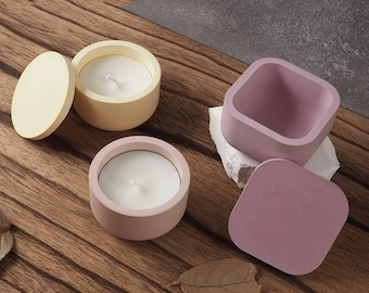 Puede incluir: Una colección de pequeños portavelas de hormigón de colores. Dos soportes redondos con velas y tapas en amarillo pastel y rosa, y un soporte cuadrado con una tapa en morado apagado. Los artículos están dispuestos sobre una superficie de madera.
