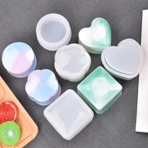May include: Set of seven silicone molds for making resin jewelry boxes. The molds are in various shapes, including round, square, heart, and flower. The molds are white and translucent, and some have a light green or pink tint.