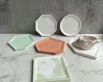 Può includere: Set di sei vassoi decorativi di diverse forme e colori, tra cui bianco, verde, arancione e verde chiaro. I vassoi sono realizzati in cemento e hanno una finitura liscia e lucida.