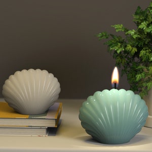 May include: Two seashell shaped candles, one is lit and is a light blue color, the other is unlit and is a light gray color.