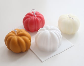 Peut inclure: Quatre petites bougies en forme de citrouille de couleur rouge, blanche, orange et crème. Les bougies sont sur une surface blanche.