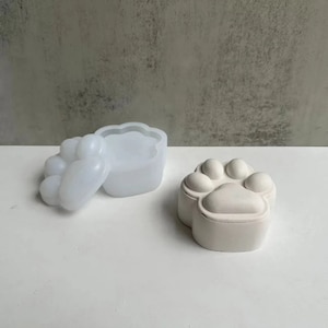 May include: A white paw print shaped mold and a white paw print shaped container. The mold is open and the container is closed.