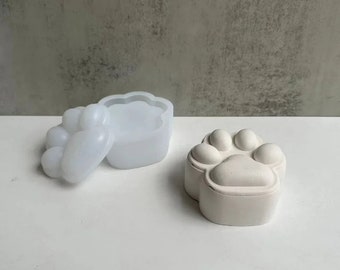 Può includere: Uno stampo bianco a forma di impronta di zampa e un contenitore bianco a forma di impronta di zampa. Lo stampo è aperto e il contenitore è chiuso.