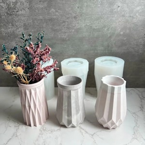 Peut inclure: Quatre vases en béton géométriques, deux de couleur rose clair et deux de couleur gris clair. Les vases roses sont remplis de fleurs séchées.