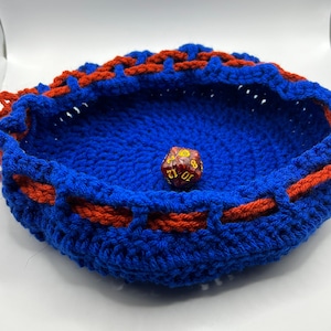 May include: A blue and red crocheted dice rolling tray with a red and gold dice in the center.