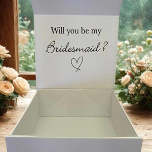 May include: A white rectangular gift box with a hinged lid that stands open. The interior of the lid features the black printed text "Will you be my Bridesmaid?" followed by a small hand-drawn heart symbol centred below the question.