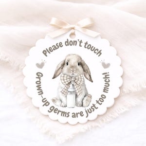 May include: A white, scalloped-edge tag featuring a watercolour illustration of a bunny with a plaid bow tie and a bottle. The tag says "Please don't touch! Grown-up germs are just too much!" A cream ribbon is attached.