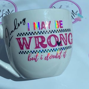 May include: White ceramic mug with the text "I MAY BE WRONG but i doubt it" in pink and holographic lettering. The mug also has the name "Lindsey" in cursive. The mug has a handle and is on a white surface.