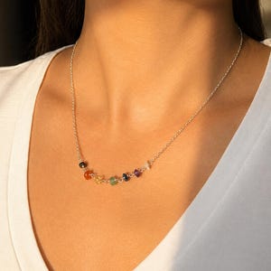 May include: A silver necklace with a fine chain, adorned with seven multi-coloured gemstone beads. The gemstones are arranged in a line, displaying a range of colours: black, orange, yellow, green, blue, purple, and clear.