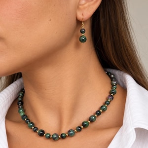 May include: A necklace and a single earring featuring round, dark green and speckled gemstone beads interspersed with small, polished gold-tone metal spacers. The necklace consists of a single strand of beads, while the earring has two beads.