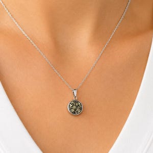 May include: A silver necklace with a circular pendant. The pendant is made up of small, dark green and gold-coloured stones. The necklace is worn against a white v-neck top.