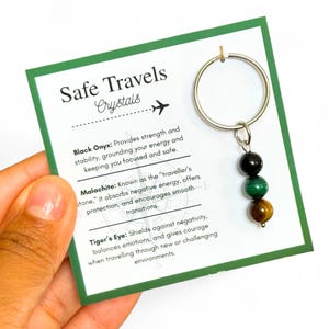 May include: A keychain with three gemstone beads: black onyx, malachite, and tiger's eye. The keychain is attached to a green card with the text "Safe Travels Crystals" and descriptions of each stone's properties.