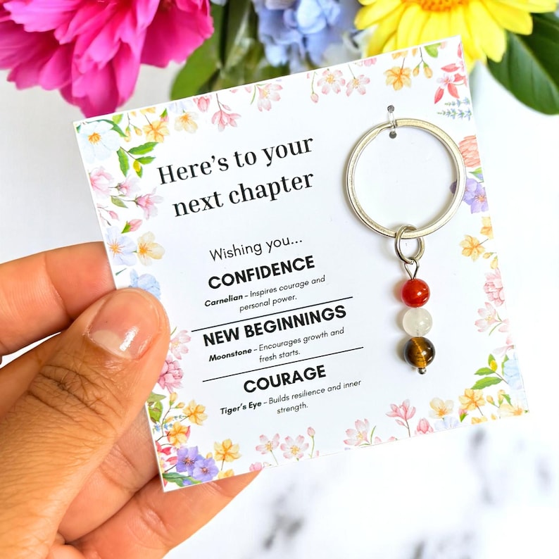 May include: A keychain with a silver ring and three gemstone beads: red, white, and brown. The keychain hangs from a card with floral accents and the text "Here's to your next chapter". The card also lists "Confidence", "New Beginnings", and "Courage".