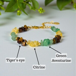 Puede incluir: Una pulsera dorada con una variedad de piedras preciosas. La pulsera incluye ojo de tigre, citrino y aventurina verde. Las piedras preciosas están dispuestas en un patrón a lo largo de la pulsera.
