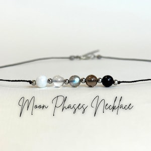 Puede incluir: Un collar de cordón negro con cinco cuentas de piedras preciosas en blanco, transparente, iridiscente, marrón y negro. El collar tiene pequeñas cuentas de color plateado. El texto "Moon Phases Necklace" está escrito en cursiva debajo.