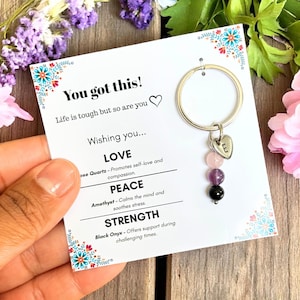 Puede incluir: Un llavero plateado con un dije en forma de corazón grabado con la letra "E" y tres cuentas de piedras preciosas: rosa, morado y negro. El llavero cuelga de una tarjeta con el texto "You got this!" y "Wishing you... LOVE, PEACE, STRENGTH."