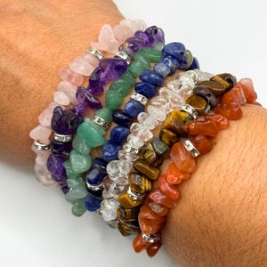 May include: A stack of seven bracelets made with different colored semi-precious stones. The bracelets are made with chips of rose quartz, amethyst, green aventurine, lapis lazuli, clear quartz, tiger's eye, and carnelian.