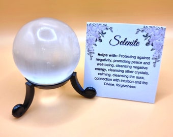 Puede incluir: Una esfera de selenita transparente sobre un soporte negro, con una tarjeta que dice "Selenita". La tarjeta enumera los beneficios de la selenita, incluyendo la promoción de la paz y la limpieza de la energía negativa. La esfera es lisa y translúcida.