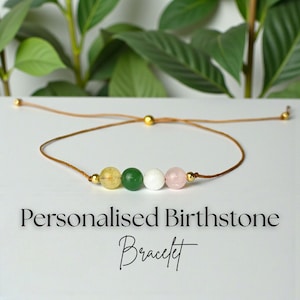 Puede incluir: Una pulsera de piedras de nacimiento personalizada con cuatro cuentas de piedras preciosas de colores: amarillo, verde, blanco y rosa. Las cuentas están ensartadas en un cordón marrón con detalles dorados. El texto "Personalised Birthstone Bracelet" está debajo.
