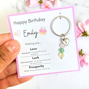 以下が含まれることがあります： ピンク色のカードが付いた誕生日をテーマにしたキーホルダー。「Happy Birthday Emily」と「Wishing you... Love, Luck, Prosperity」と書かれています。キーホルダーには、文字「S」のハートチャームと、ローズクォーツ、グリーンアベンチュリン、シトリンの3つの宝石ビーズが付いています。