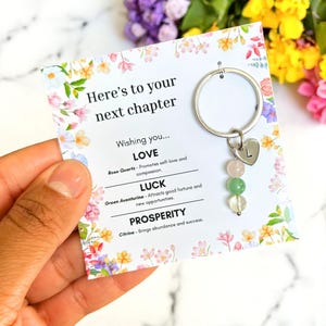 Könnte beinhalten: Ein Schlüsselanhänger mit einem silbernen Ring und einem herzförmigen Anhänger mit dem Buchstaben "L". Darunter befinden sich drei Edelsteinperlen: rosa, grün und klar. Der Schlüsselanhänger ist an einer Karte mit floralen Rändern und dem Text "Here's to your next chapter" befestigt.