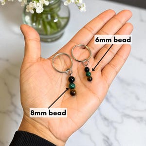 May include: Two keychains with silver rings, each featuring a series of beads in shades of black, green, and brown. One keychain has 8mm beads, the other has 6mm beads. The keychains are displayed on a hand.