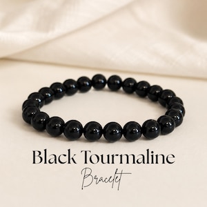 May include: A stretch bracelet composed of smooth, round, polished black tourmaline beads. The text "Black Tourmaline Bracelet" is printed in black lettering below the jewelry piece, which is arranged in an oval shape on a light surface.