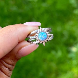 May include: A silver ring set featuring a starburst design with a light blue opal centre stone. The ring has a twisted rope band. The ring is held up against a blurred green background.