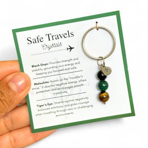 May include: A keychain with a silver ring and a charm with the letter "M" in a heart shape. The keychain has three gemstone beads: black onyx, malachite, and tiger's eye. The card reads "Safe Travels Crystals."