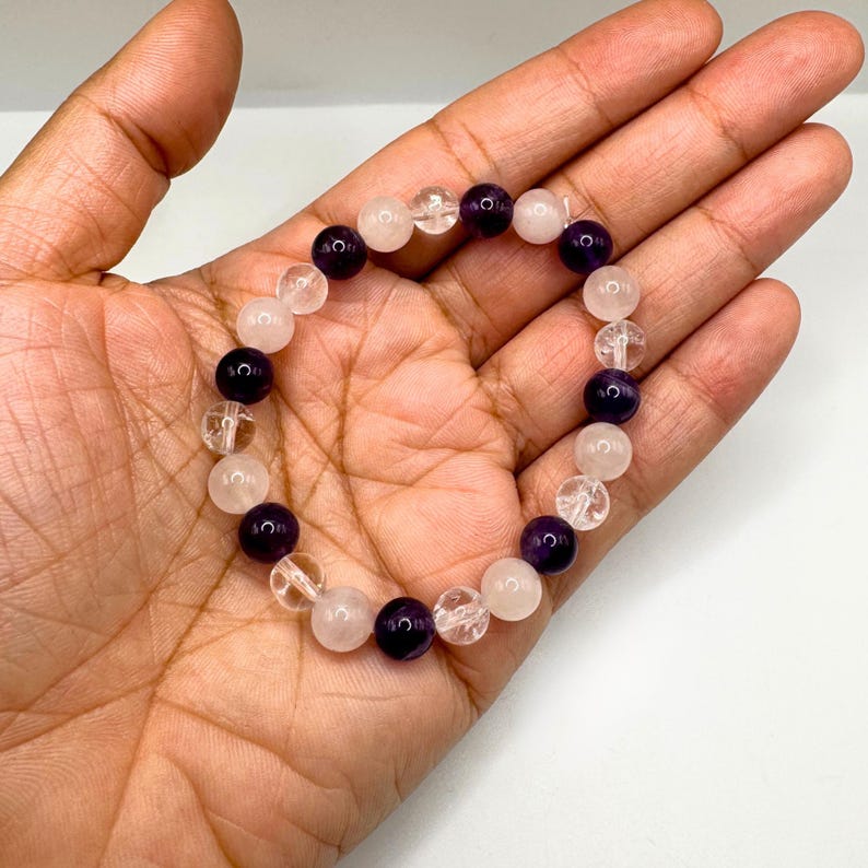 May include: A bracelet made with clear, pink, and purple beads. The bracelet is on a person's hand.