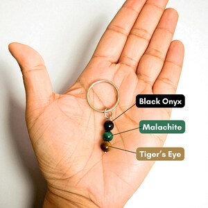 May include: A keychain featuring three gemstone beads: black onyx, malachite, and tiger's eye. The beads are suspended from a gold-colored ring. The names of the gemstones are labeled in the image.