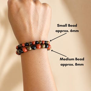 K&ouml;nnte beinhalten: Zwei Armb&auml;nder mit Perlen, kleine Perlen von ca. 6 mm und mittlere Perlen von ca. 8 mm. Die Armb&auml;nder haben eine Mischung aus roten, schwarzen und goldfarbenen Perlen. Die Armb&auml;nder werden an einem Handgelenk getragen.