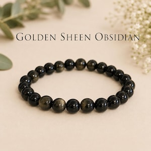May include: A stretch bracelet composed of round, polished beads made of golden sheen obsidian. The beads are primarily black with metallic gold inclusions that catch the light. The text "GOLDEN SHEEN OBSIDIAN" is printed above the bracelet.