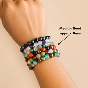 K&ouml;nnte beinhalten: Eine Sammlung von Armb&auml;ndern mit Perlen in verschiedenen Farben, darunter Schwarz, Lila, Rot, Gr&uuml;n und Gelb. Die Perlen sind etwa 8 mm gro&szlig;. Die Armb&auml;nder sind &uuml;bereinander gestapelt.
