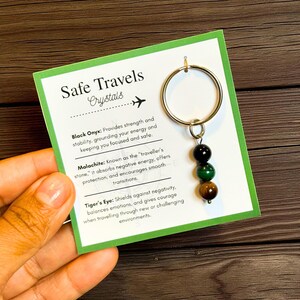 May include: A gold-colored keychain with three gemstone beads: black onyx, green malachite, and tiger's eye. The keychain hangs from a card with the text "Safe Travels Crystals" and descriptions of each crystal's properties.