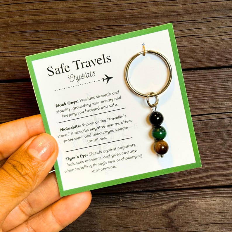 May include: A gold-colored keychain with three gemstone beads: black onyx, green malachite, and tiger's eye. The keychain hangs from a card with the text "Safe Travels Crystals" and descriptions of each crystal's properties.