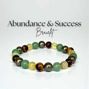 May include: A bracelet made of various coloured gemstones, including green, yellow, brown, and silver beads. The bracelet is displayed on a white surface. Text above the bracelet reads "Abundance & Success Bracelet."