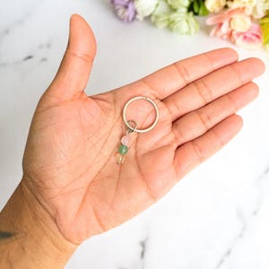 May include: A silver-colored keychain with three small, colorful gemstone beads: pink, green, and clear. The keychain is held in an open hand, with a marble-like surface in the background. The beads are round and smooth.