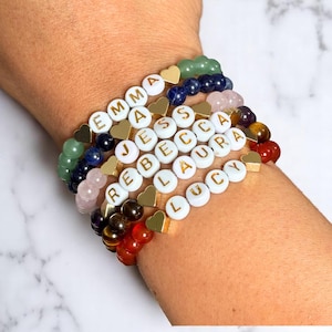 Puede incluir: Un conjunto de siete pulseras con cuentas blancas y dijes de corazón dorados. Cada cuenta tiene una letra que deletrea los nombres Emma, Jessica, Rebecca, Laura y Lucy. Las pulseras están hechas con piedras preciosas de diferentes colores, incluyendo verde, azul, rosa, marrón y rojo.